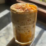 A close-up view of a creamy Peach Cobbler Latte in a clear glass, topped with whipped cream and sprinkled with cinnamon.