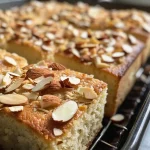 Close-up of a moist almond vanilla snack cake with a golden crust and sliced almonds on top.