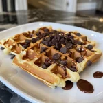 A close-up side view of golden brown chocolate chip waffles stacked on a plate.