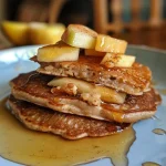 ['Close-up view of stacked Apple Cinnamon Protein Pancakes on a plate.', 'Side view of a fluffy stack of pancakes with apple and cinnamon toppings.', 'Appetizing image of protein pancakes garnished with diced apples and cinnamon.', 'Delicious Apple Cinnamon Protein Pancakes served on a wooden table.']