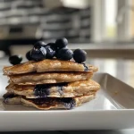 ['Close-up of a stack of golden-brown blueberry oat pancakes with fresh blueberries on top.', 'Side view of fluffy blueberry oat pancakes, drizzled with syrup and garnished with blueberries.', 'Delicious blueberry oat pancakes served on a white plate, showcasing their texture and color.', 'Juicy blueberry oat pancakes complemented by fresh fruit and a hint of syrup.']