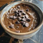['Close-up view of a creamy brown dip made from healthy ingredients in a bowl, garnished with chocolate chips.', 'Side view of a thick brownie batter dip in a white bowl, with chocolate chips scattered on top.', 'A delicious-looking vegan brownie batter dip displayed in a bowl with a smooth texture and chocolate sprinkles.', 'Close-up of a healthy brownie batter dip in a bowl, emphasizing its rich brown color and creamy consistency.']