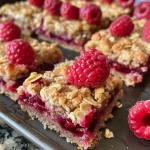 ['Close-up view of raspberry bars with oats and a crumbly texture.', 'Side view of freshly made raspberry bars, showing layers of fruit and dough.', 'Juicy raspberry bars with a golden crust, highlighted by a soft focus background.', 'Delicious raspberry dessert bars, emphasizing the vibrant red fruit filling.']