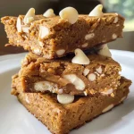 ['Close-up side view of a delicious White Chocolate Pumpkin Protein Blondie with a golden-brown crust.', 'A moist and inviting White Chocolate Pumpkin Protein Blondie, showcasing white chocolate chips and a textured surface.', 'A vibrant pumpkin protein blondie highlighted by chunks of white chocolate, arranged on a white plate.', 'A side view of a gluten-free White Chocolate Pumpkin Protein Blondie with visible spices and creamy texture.']