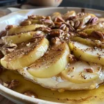 ['Close-up of a baked brie topped with apple slices and pecans.', 'Side view of gooey baked brie with caramelized apples and honey drizzle.', 'Juicy baked brie adorned with fresh apple slices and a sprinkle of brown sugar.', 'Melted brie cheese with apple wedges and pecans, presented on a wooden board.']