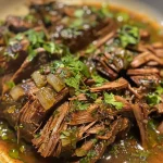 ['Close-up of juicy Beef Barbacoa in a bowl, garnished with cilantro.', 'Side view of tender Beef Barbacoa, showcasing its rich color and texture.', 'Detailed image of slow-cooked Beef Barbacoa with lime wedges and cilantro.', 'Appetizing display of Beef Barbacoa served in a rustic bowl.']