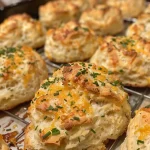 ['Close-up view of golden-brown Cheddar Chive Drop Biscuits on a rustic wooden table.', 'Side view of freshly baked Cheddar Chive Drop Biscuits, showcasing their flaky texture and cheesy interior.', 'Juicy Cheddar Chive Drop Biscuits piled on a white plate, garnished with fresh chives.', 'Delicious Cheddar Chive Drop Biscuits displayed against a dark background, capturing their buttery shine.']