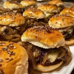 ['Close-up view of French onion beef sliders stacked on a serving platter.', 'Juicy French onion beef sliders with melted cheese and sautéed onions.', 'Side view of delicious beef sliders topped with gruyere cheese and onions.', 'A tempting arrangement of French onion beef sliders ready to serve.']