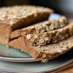 ['Side view of a loaf of healthy vegan oatmeal bread, showcasing a golden-brown crust.', 'Close-up of freshly baked vegan oatmeal bread with visible oats on the surface.', 'Slice of healthy vegan oatmeal bread, highlighting its texture and color.', 'Whole loaf of vegan oatmeal bread positioned on a rustic wooden table.']