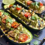['Close-up view of Pesto Chicken Stuffed Zucchini Boats with vibrant toppings.', 'Juicy zucchini boats filled with pesto chicken, garnished with cherry tomatoes and cilantro.', 'Side view of baked zucchini halves stuffed with chicken, melted mozzarella, and pesto sauce.', 'Deliciously presented zucchini boats topped with cheese and fresh ingredients.']