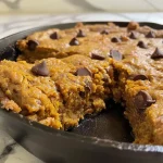 ['A close-up side view of a freshly baked Pumpkin Skillet Cookie with dark chocolate chips.', 'A warm, golden-brown Pumpkin Skillet Cookie served in a cast iron skillet, showcasing its gooey texture.', 'A delicious Pumpkin Skillet Cookie with melting chocolate chips, set against a rustic kitchen background.', 'A side view of a Pumpkin Skillet Cookie, highlighting its soft, inviting appearance and rich ingredients.']