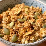 ['Close-up view of Pumpkin Spice Protein Granola in a bowl, highlighting pumpkin seeds and oats.', 'Side angle shot of crunchy Pumpkin Spice Protein Granola with spices and seeds visible.', 'Textured granola mix featuring pumpkin spices, oats, and seeds in a bowl.', 'Detailed close-up of golden-brown Pumpkin Spice Protein Granola with ingredients showing.']