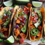 ['Close-up view of roasted sweet potato tacos with avocado lime crema and fresh toppings.', 'Side view of juicy roasted sweet potato tacos garnished with avocado crema and colorful vegetables.', 'Delicious roasted sweet potato tacos featuring black beans, cilantro, and lime wedges on a plate.', 'A vibrant arrangement of sweet potato tacos topped with salsa, jalapeño, and creamy avocado dressing.']