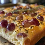 ['A side view of a golden-brown focaccia topped with juicy red grapes and fresh rosemary.', 'Close-up of rosemary and grape focaccia, showcasing grapes nestled in the bread’s crust.', 'Freshly baked focaccia with a crispy exterior, adorned with rosemary and clusters of red grapes.', 'A vibrant focaccia bread highlighting red grapes and rosemary sprigs on its surface.']
