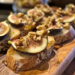 ['Close-up view of a fig and walnut crostini topped with cheese and honey.', 'Side view of crostini with fig jam, walnuts, and cheese, drizzled with honey.', 'Appetizing fig and walnut crostini on a wooden board, showcasing vibrant toppings.', 'Delicious crostini featuring fig jam, chopped walnuts, and Iberico cheese with a honey drizzle.']